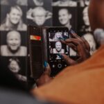 A person holds a smartphone in a brown wallet case, taking a photo of a black-and-white portrait displayed on a wall filled with similar portraits. The phone's screen shows the captured image, while another person partially appears in the foreground.