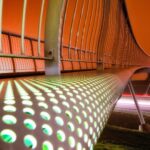 A modern bridge with an illuminated perforated side rail is captured at night. The long exposure results in a dramatic effect, with trailing lights from passing vehicles below. The sky is a warm, glowing orange, enhancing the futuristic appearance of the bridge.