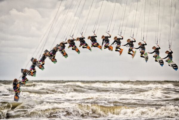 A sequence of images shows a kiteboarder performing a jump and trick over rough, choppy ocean waves, forming an arc in the sky. The cloudy sky and turbulent sea emphasize the dynamic and intense nature of the sport.