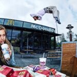 A young woman with long hair sips a drink and eats French fries at an outdoor McDonald's seating area. In the background, a man is captured mid-air performing an impressive flip. Other people sit nearby, and the McDonald's building with its logo is clearly visible.