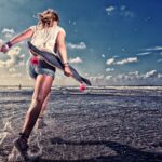 A person with wet legs and a skateboard held behind their back is running towards the ocean under a partly cloudy sky. The waves are gently rolling in, and the beach is reflecting the light, creating a dynamic and vibrant scene.