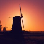 Silhouettes of three traditional windmills stand against a vivid orange and purple sunset sky. The scene is tranquil, with an open field in the foreground and a line of trees in the background, capturing the serene essence of the countryside.