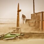 A person stands on a beach next to an orange building in a windy and sandy environment. A small green boat is partially buried in the sand nearby, and visibility is low due to the blowing sand. The scene appears desolate and stormy.