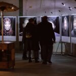 A group of people stand in front of a series of large black-and-white portrait photographs displayed on a wall in an indoor art gallery. The portraits are illuminated, and the viewers are engaged, observing the details of the artwork. A heater stands nearby.