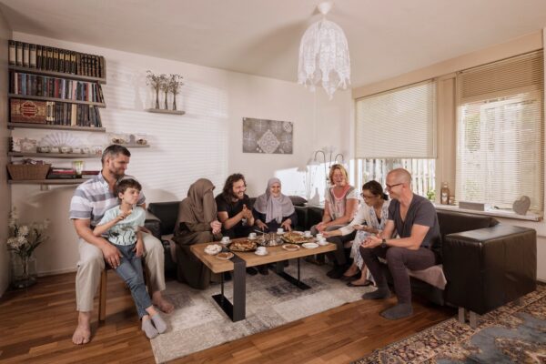 A group of people, including adults and children, are sitting around a low table in a cozy living room. They are sharing a meal and appear to be engaged in conversation. The room has modern decor and large windows with blinds partially open.