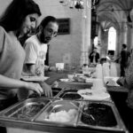 Black and white photo of three people standing at a buffet table in a large hall with arched ceilings and chandeliers. One woman is serving herself food, a man is holding a plate and bowl, and another woman, wearing a headscarf, is also serving food.