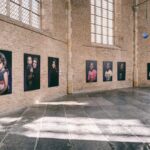 A spacious room with large windows, featuring a brick interior and high ceilings. Six large portrait photographs are displayed on the walls, showcasing diverse individuals. Sunlight streams through the windows, casting patterns on the floor. A door is visible in the back.