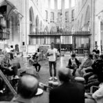 A black and white image of a diverse group of people seated in a circle inside a large, ornate church. One person stands in the center, speaking or singing, while others play various musical instruments such as guitars and drums. The atmosphere appears communal and artistic.