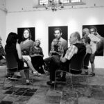 A group of men and women sit in a circle inside an art gallery, engaged in a discussion. Behind them hang large, black-and-white portrait photographs on white walls. The room has tiled flooring and a chandelier hanging from the ceiling.