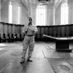 A person in military attire stands in the center of a historic, cathedral-like room with tall, arched windows. The individual wears a cap and sunglasses and holds a camera. The room features a large touchscreen display and ornate wooden paneling on the walls.