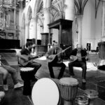 A group of four men are seated in a semi-circle inside a large, ornate hall with high vaulted ceilings. Three of them are playing stringed instruments, while the fourth man listens. Various drums and percussion instruments are placed on the floor around them.