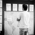 A person with short, curly hair stands with arms crossed, viewing a large board displaying a world map. Papers with text are pinned around the map, each connected to specific locations with string. The setting appears to be indoors.