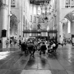 A black and white photo captures the interior of a large, historic church with high ceilings, ornate chandeliers, and tall pillars. A group of people is seated in a circle in the center, while others are scattered around the space, engaged in various activities.