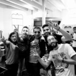 A black-and-white photo shows a group of six smiling people standing close together in what appears to be a kitchen. Several individuals are making peace signs with their hands, while one person leans forward noticeably, holding a phone. The background reveals kitchen equipment.