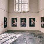 A gallery room with high ceilings and an antique chandelier features five portrait photographs displayed on a light-colored wall. The portraits depict individuals with varying expressions. Sunlight filters through tall, arched windows, casting shadows on the stone floor.