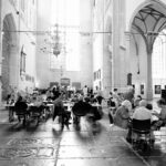 A group of people sit at round tables inside a grand, high-ceilinged hall with arched columns and large windows. The black-and-white image captures the space's intricate architectural details and the people engaged in conversation or activities.