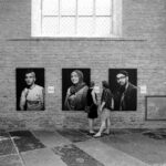 Two women stand closely together in front of three large portrait photographs hanging on a brick wall inside a building with high windows. The portraits depict different individuals, and the setting appears to be an art exhibition or gallery. The scene is in black and white.