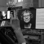 A person takes a photo with their smartphone of a large black-and-white portrait displayed on an easel in a gallery. The portrait features an older woman with curly hair and glasses. There is descriptive text about the portrait on a piece of paper next to it.