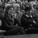 A black and white photo showing a diverse group of people sitting closely together in an audience. In the forefront, a woman and a man sit with their arms crossed, attentively watching something off-camera. Other audience members appear engaged and reactive.