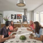 Four people are gathered around a dining table in a modern, white kitchen. They are smiling and eating a meal together. The table is decorated with a patterned tablecloth and a centerpiece. Behind them, there is kitchen equipment, art, a large window, and sliding glass doors.