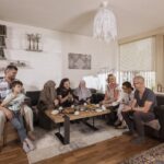 A diverse group of people sits around a low square table, enjoying a meal together in a cozy living room. They appear to be engaged in conversation and laughter. The room is decorated with bookshelves, wall art, and soft lighting, creating a warm atmosphere.