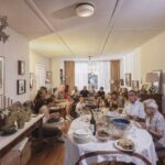 A lively family gathering in a cozy living room. Several people of various ages are seated around, sharing a meal. The room is adorned with framed pictures, a chandelier, and a side table with decor. A dining table with food and drinks is in the foreground.