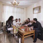 Five people are gathered around a wooden table in a living room, sharing a meal that includes a tagine and a variety of fruits. The room is warmly lit, with a sofa, a large trophy on a side table, and a chandelier overhead. Everyone appears to be enjoying the meal.