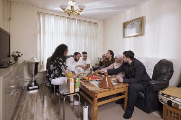 Five people are gathered around a wooden table in a living room, sharing a meal that includes a tagine and a variety of fruits. The room is warmly lit, with a sofa, a large trophy on a side table, and a chandelier overhead. Everyone appears to be enjoying the meal.