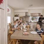 A family with four children sits at a wooden dining table enjoying breakfast in a cozy kitchen. The table is set with milk, cereal, toast, and other breakfast items. An adult is standing near the stove preparing more food. Children's drawings are visible on the wall.