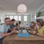 A family of four sits around a wooden dining table in a modern, brightly lit room with light green walls. They are engaged in conversation while enjoying a meal together. The room is decorated with plants and minimalistic artwork. A white pendant lamp hangs overhead.