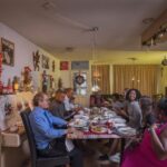 A large family gathers around a long dining table, sharing a meal in a warmly decorated room. Holiday decorations, including tinsel and garlands, are visible on the walls and furniture. Everyone appears engaged in conversation, creating a festive, joyful atmosphere.