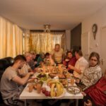 A group of people are gathered around a dining table sharing a meal in a warmly lit room. The table is filled with various dishes, drinks, and a fruit basket. They appear to be engaged in conversation and enjoying the meal together. The atmosphere is cozy and intimate.