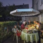 A family is gathered around an outdoor table for a meal in the evening. They are sitting under a large white umbrella, with various dishes on the table covered with a green and white checkered tablecloth. Three children and five adults are present, all engaged in conversation.