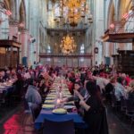 A large group of people is seated at long, decorated tables inside a grand church with high vaulted ceilings and chandeliers. The tables are set with blue tablecloths, plates, and glasses, and attendees are engaged in conversation. The atmosphere is festive and elegant.