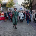 A group of people follows a man in a green robe walking down a brick-paved street in a city. The crowd includes men and women of various ages, some of whom are smiling. Buildings and a colorful, partly visible structure are in the background.