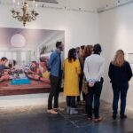 A group of people wearing headphones stand in an art gallery, closely examining a large photograph on the wall. The photograph depicts a family dining together around a wooden table. A red fire hose and an informational plaque are visible on the right wall.