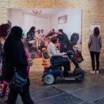 A diverse group of people in a gallery observe a large photograph displayed on a brick wall. The photo features a man on a mobility scooter, a woman with dreadlocks in a floral dress and others. The setting has a contemporary, inclusive atmosphere.