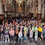 A large, diverse group of people are gathered inside a spacious hall with high ceilings, pillars, and chandeliers. They are smiling and looking at the camera. Some individuals are seated, while most are standing, with children in the front.