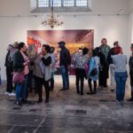 A group of people stands in an art gallery, many wearing headphones. They gather around a large, vibrant photograph displayed on a white wall. The space has high ceilings, a chandelier, stone flooring, and large windows, creating a bright and airy atmosphere.