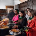 A group of people with green headphones wait in line to be served soup from large metal pots in a buffet-style setting. They are indoors, in an area with dark wooden paneling. Some hold bowls, and a person ladles soup into a bowl.