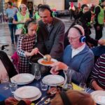 A group of people of various ages, all wearing wireless headphones, gather around a table set with white plates and glasses. A young girl serves soup from a bowl to an older man seated across from her. The mood is communal and lively.