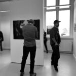 A black and white photo inside an art gallery. Several people observe artwork on display. A man in the foreground faces a large portrait close-up of a woman's face, while another man with a cap stands nearby looking at other pieces. Large windows let in natural light.