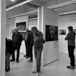 A black and white photo shows people in an art gallery viewing various photographic artworks. One man stands close, looking intently at a portrait piece. Others are dispersed throughout the room, all seemingly engaged with the exhibition.