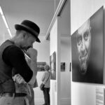 A man wearing a hat, suspenders, and casual attire stands pensively in an art gallery, observing a large photograph of a face. Other gallery visitors are visible in the background, some seated and others examining the artwork. Black and white photo.