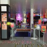 Entrance to an arcade with a brightly lit hallway leading to gaming areas. Balloons are tied to displays on either side of the entrance. Posters and banners are visible, advertising arcade attractions. Lights and decorations create a festive atmosphere.