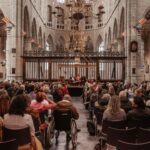 A large audience seated inside a historic stone cathedral, focusing on a stage where several speakers are seated at a table. The cathedral features high arched ceilings, ornate chandeliers, and intricate wooden and metal designs. A person in a wheelchair is in the foreground.