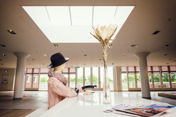 A mannequin dressed in a pink coat, black hat, and scarf, holding a cup sits at a white counter in a spacious, modern room with tall windows. A tall vase with a single large flower is on the counter, and a couple of magazines are placed to the right.