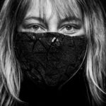 Black and white close-up of a person with long hair wearing a black sequin face mask. The mask covers their nose and mouth, and their eyes look directly at the camera. The background is dark, focusing attention on the person's face.