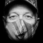 Black and white close-up of a person wearing a cap and a striped cloth face mask. The person's eyes are looking directly into the camera. The background is dark, making the face stand out prominently.