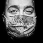 Black and white close-up of a person wearing a floral-patterned face mask. Their expressive eyes are wide open and focused, while the mask covers the lower half of the face. The background is dark, highlighting the subject's features and the texture of the mask.